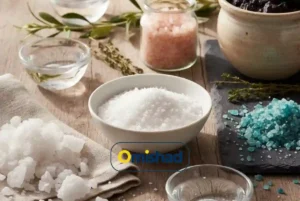 نمک سفید، صورتی و آبی امیشاد در ظروف شیشه‌ای و سرامیکی روی میز چوبی روشن با نور طبیعی و گیاهان تزئینی.