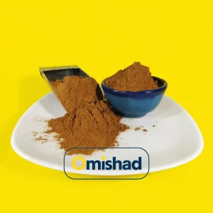 توده پودر دارچین قهوه‌ای تیره با بافت نرم در کاسه سفالی آبی و پیمانه استیل بر روی بشقاب سفید.
