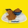 پودر دارچین ریز و خوش‌رنگ در کاسه سرامیکی آبی و روی بشقاب سفید کنار پیمانه فلزی با زمینه زرد.
