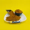 توده ادویه کاری پودری با تم قهوه‌ای-نارنجی در مرکز بشقاب سفید، همراه با کاسه آبی لب‌پر و بیلچه فلزی استیل در زمینه زرد یکدست.