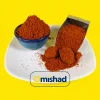 نمای نزدیک از پودر پاپریکای قرمز و خوشرنگ در کاسه سفالی آبی و پیمانه استیل روی دیس سفید؛ نمایی جذاب از ادویه با کیفیت و یکنواخت میشاد.