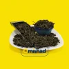 نمای نزدیک از برگهای پهن و خشکشده چای سبز ایرانی در ظرف سفالی و اسکوپ فلزی، نمایشگر خلوص و فرآوری طبیعی چای داخلی در پسزمینه زرد میشاد.
