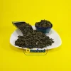 تصویر هنری برگهای خشک و پیچخورده چای سبز خارجی اعلا در پیمانه فلزی و کاسه فیروزهای روی بشقاب سفید با پسزمینه زرد و لوگوی میشاد، نماد کیفیت چای فله.