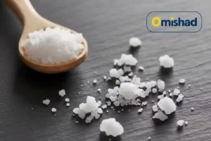 دانه‌های درشت نمک فلوریدی امیشاد پخش‌شده روی سطح تیره همراه با قاشق چوبی پر از کریستال‌های سفید.