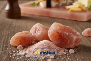 بلورهای درشت نمک صورتی هیمالیا روی میز چوبی در کنار تخته سنگ نمکی و آسیاب چوبی.